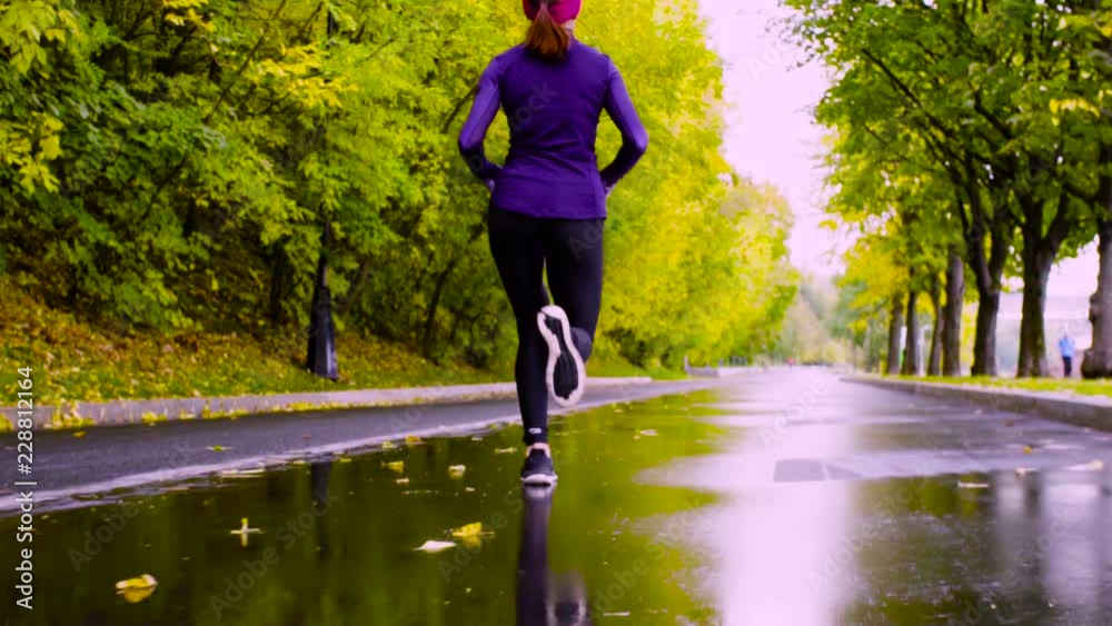 Dolly shot, slow motion. Young woman running in the park at autumn day. Rear view