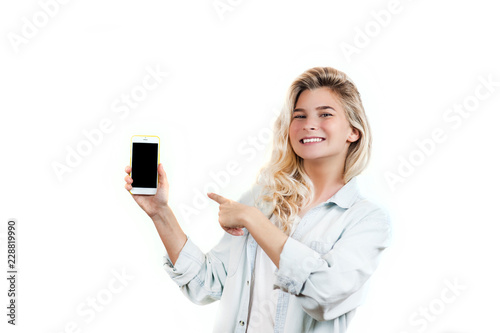very beautiful young girl points to the smartphone, standing on a white isolated background.