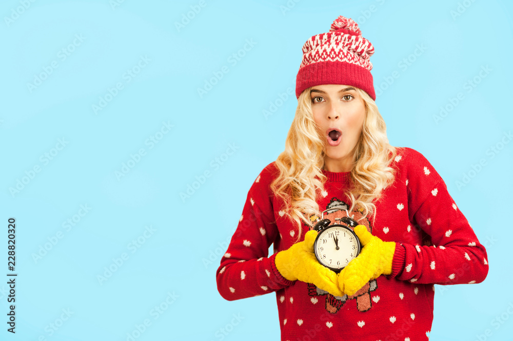 Very beautiful young bright blonde in festive Christmas clothes with a clock in her hands on a blue isolated background