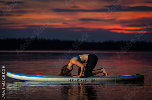 Beautiful slim fit girl with long brown hair exercising yoga on paddleboard in the dark, colorful sunset on scenic lake Velke Darko near Zdar nad Sazovou, Czech Republic photo