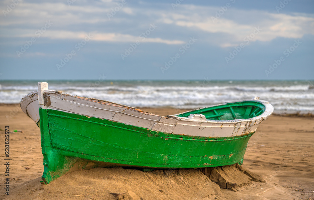 An abandoned small bright green and white wooden boat on brown sand with the waves and a blue sky and white clouds in the background