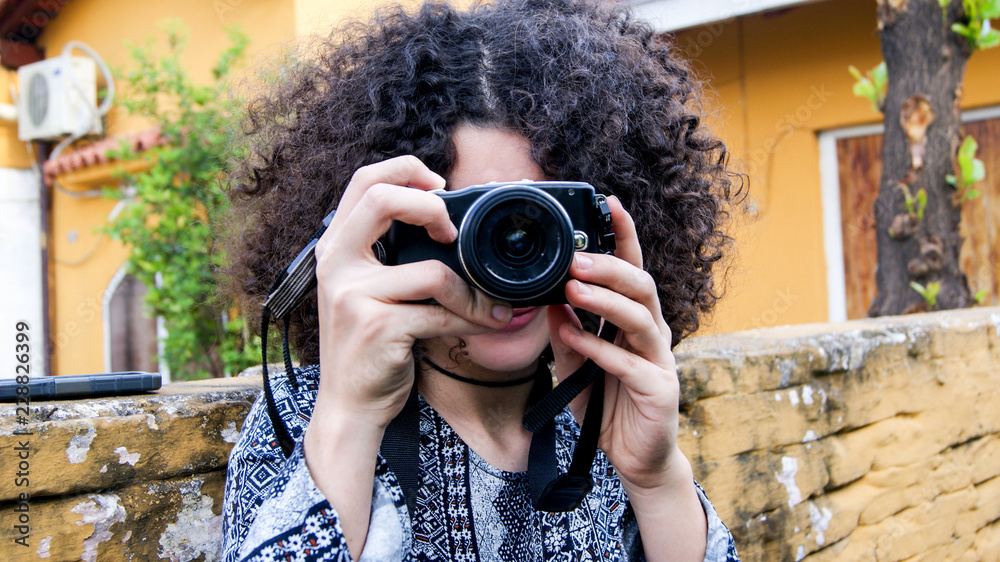 Young brunette girl with afro curly hair taking a photo with a photography proffesional camera in the street - lense looking at the camera - amateur woman photographer