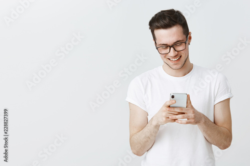 Guy texting girlfriend on way to meating spot holding smartphone in hands laughing and smiling cute as looking at device screen reacting positive and joyful to cute message posing over white wall © Cookie Studio