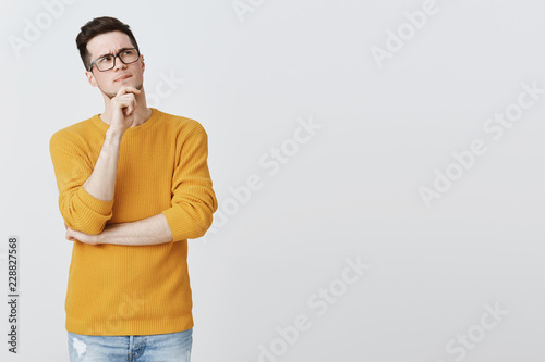 Hmm what if. Smart and thoughtful good-looking guy in geek glasses and cozy yellow sweater holding hand on chin, frowning and looking at upper right corner as thinking, making decision or assumption