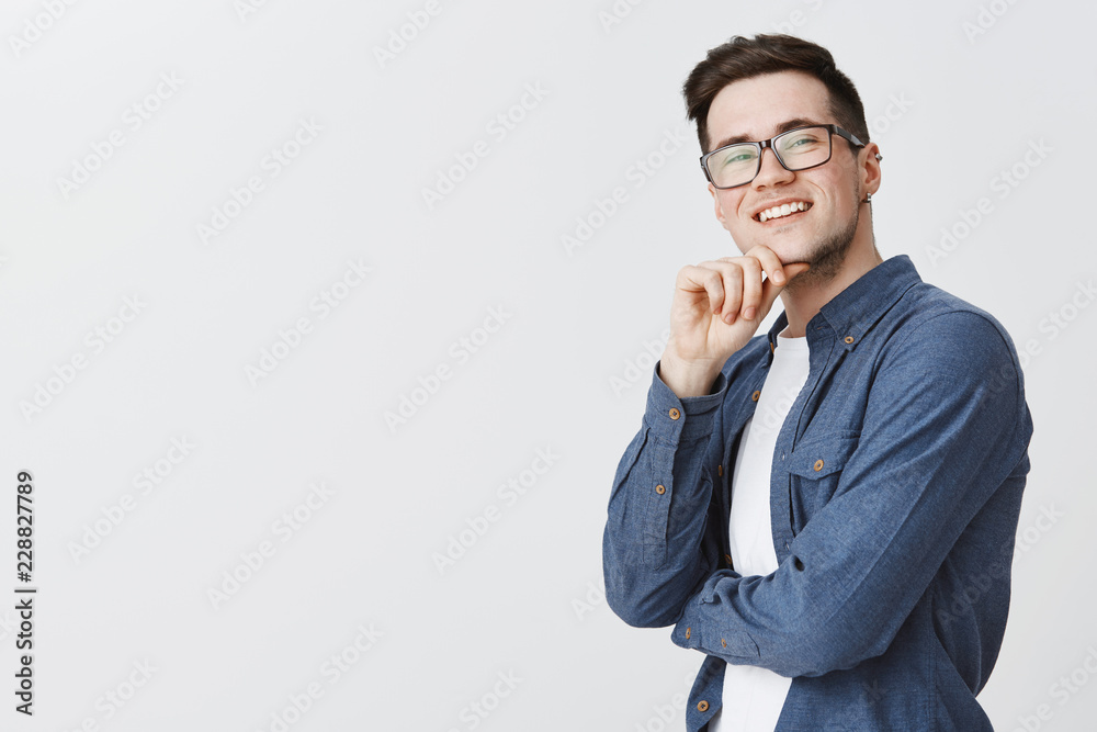 Delighted happy and satisfied handsome young male student in glasses and blue shirt holding hand on chin and smiling being satisfied with idea coming to mind, standing pleased over grey wall Delighted happy and satisfied handsome young male student in glasses and blue shirt holding hand on chin and smiling being satisfied with idea coming to mind, standing pleased over grey wall