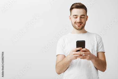 Checking profile of girl he loves. Handsome delighted and happy caucasian guy with cool haircut and bristle holding smartphone looking at device screen with satisfaction reacting to daring message © Cookie Studio