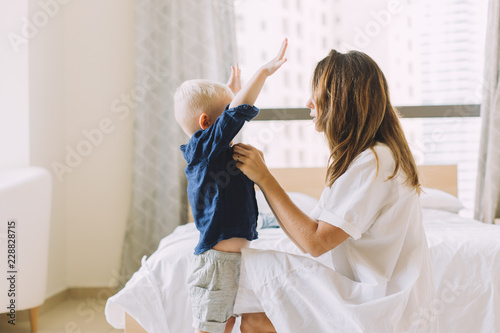 Young beautiful mother buttoning up the shirt of her little son in the bedroom photo