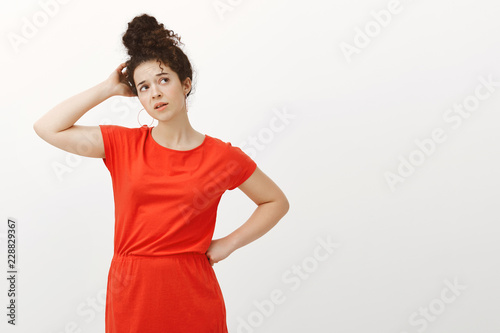 Indoor shot of confused questioned pretty female with curly hair in trendy red dress, holding hand on hip and scratching head while looking up and thinking, using imagination over gray wall