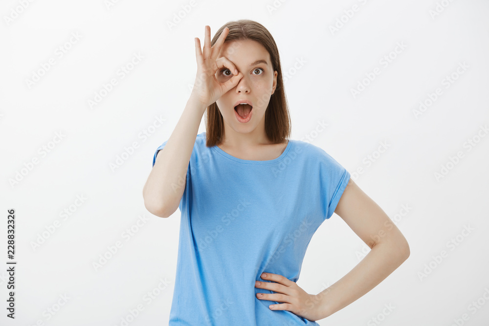 Okay, I am ready to fascinate. Good-looking playful european woman with fair hair, holding hand on hip and showing ok gesture over eye, gasping, standing with opened mouth over gray background Okay, I am ready to fascinate. Good-looking playful european woman with fair hair, holding hand on hip and showing ok gesture over eye, gasping, standing with opened mouth over gray background