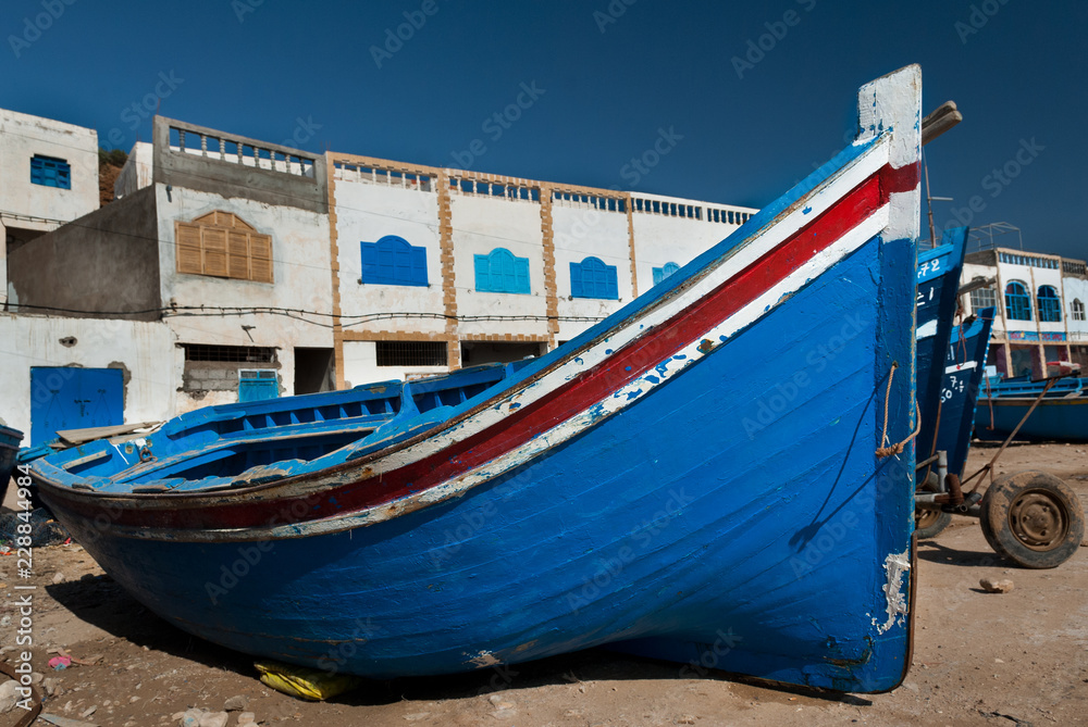 A blue boat lays on the ground in front of the Tafelney fishing village buildings