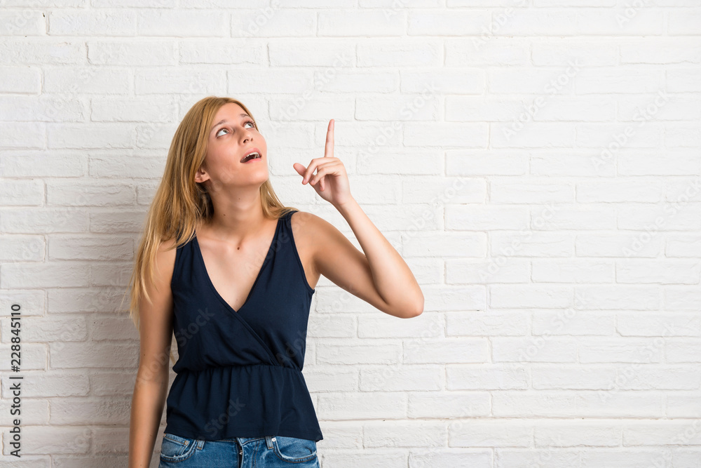 Blonde woman intending to realizes the solution while lifting a finger up on white brick wall background Blonde woman intending to realizes the solution while lifting a finger up on white brick wall background