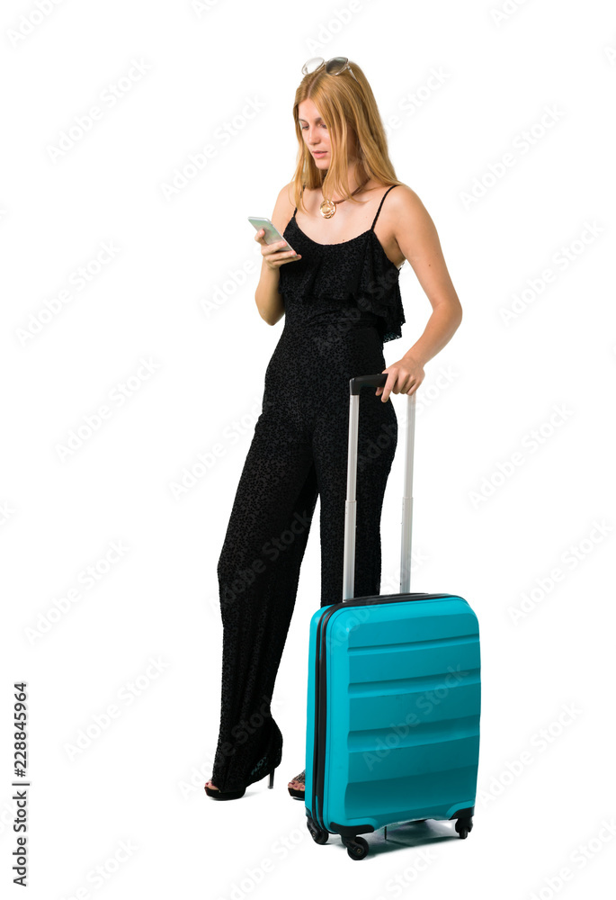 Blond girl traveling with her suitcase sending a message or email with the mobile on isolated white backgroun Blond girl traveling with her suitcase sending a message or email with the mobile on isolated white backgroun