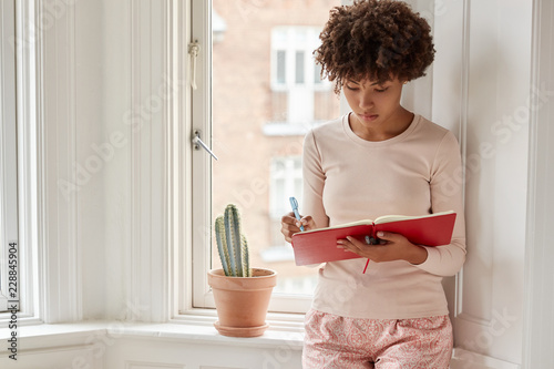 Photo of dark skinned woman has good idea at mind, writes in notepad, wears pyjamas, stands near window to have better vision and light, wears nightclothes, does list in diary against cozy interior photo
