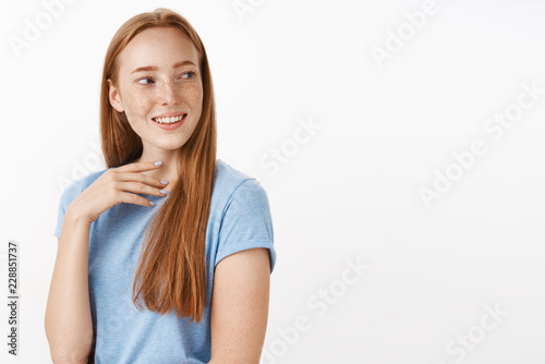 Cute beautiful young redhead woman with natural freckles turning right with broad pleased smile touching neck seeing curious and entertaining scene grinning with delight over gray wall