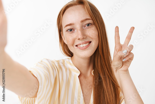 Waist-up shot of cute carefree and happy young redhead female with freckles in yellow summer dress pulling hand as if holding camera taking selfie smiling and showing peace or victory sign