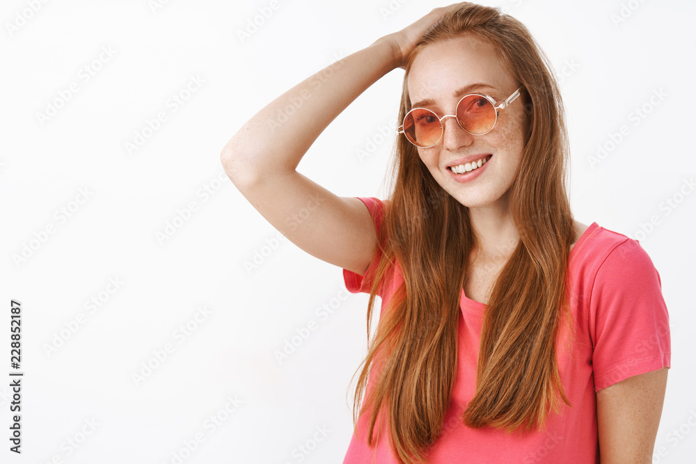 Shy and tender charming redhead lady with freckles in trendy sunglasses and pink blouse combing hair behin with hand on head and smiling friendly at camera posing over white background Shy and tender charming redhead lady with freckles in trendy sunglasses and pink blouse combing hair behin with hand on head and smiling friendly at camera posing over white background