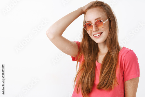 Shy and tender charming redhead lady with freckles in trendy sunglasses and pink blouse combing hair behin with hand on head and smiling friendly at camera posing over white background © Cookie Studio