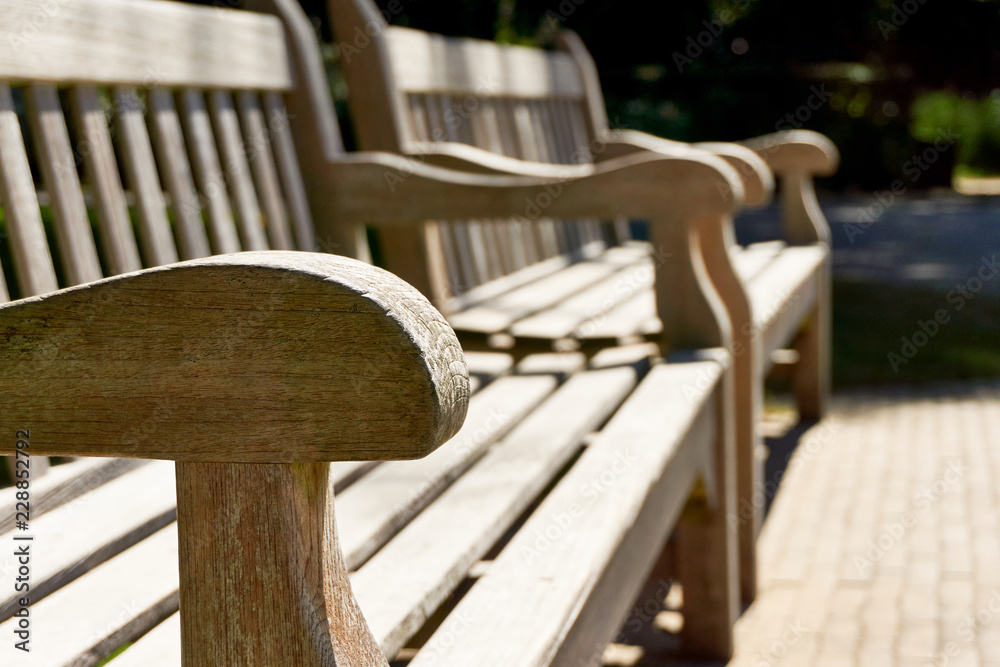 Close-up of two wooden classic teak park benches in the sun in summer with selective focus on one armrest Close-up of two wooden classic teak park benches in the sun in summer with selective focus on one armrest