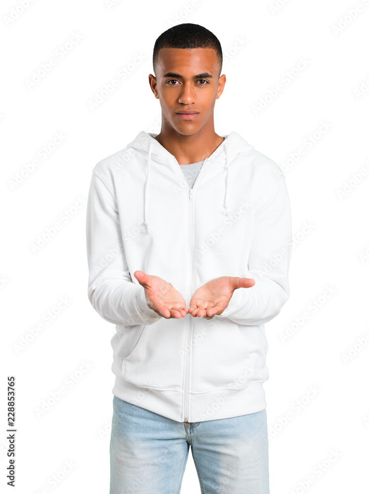 Fototapeta premium Dark-skinned young man with white sweatshirt holding copyspace imaginary on the palm to insert an ad on isolated white background