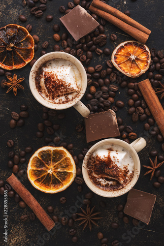 Two cups of hot chocolate with marshmallows on a rustic background decorated with coffee beans, cinnamon and orange slices. Warming winter or autumn drink. Top view. Toned photo. © Yulia
