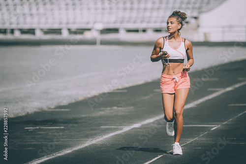 Purposeful runner female listening to running motivation music with earbuds on her mobile phone app for run on track in athletics stadium. Athlete enjoying fitness active life concept. Grey background