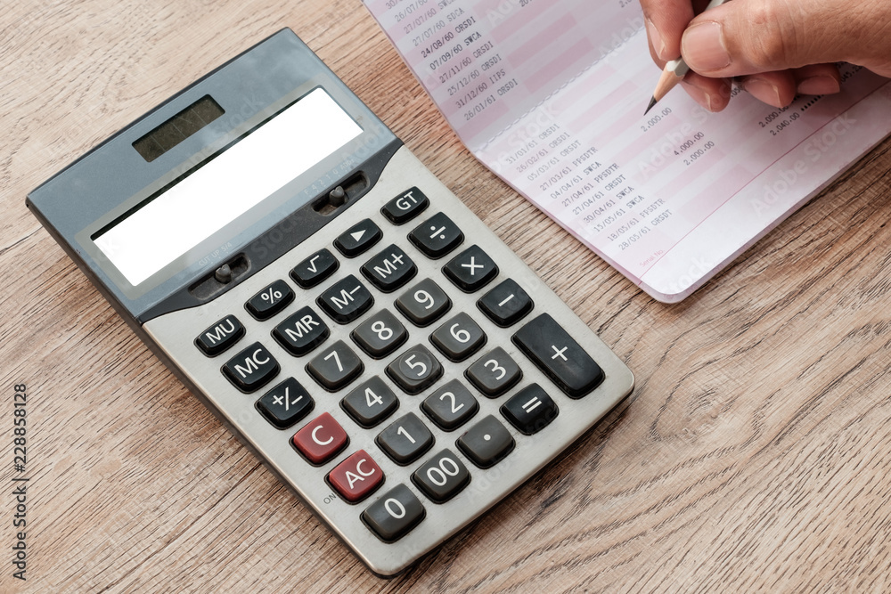 The fingers are holding the pencil. Below is a white screen calculator, a bank book, placed on a wooden desk. The fingers are holding the pencil. Below is a white screen calculator, a bank book, placed on a wooden desk.