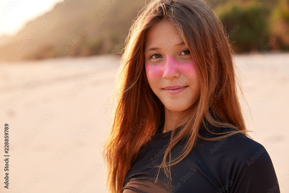 Close up shot of beautiful Caucasian girl with pink zurf zinc mask, dressed in black wetsuit, enjoys sunshine, looks happily directly at camera, has recreation time in exotic paradise country