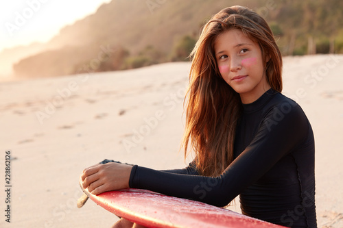 Photos Contemplative Caucasian young woman with long hair, surf zinc on cheeks, carries surfboard, looks pensively aside, poses on beach with free space for your advertising content or promotional text
