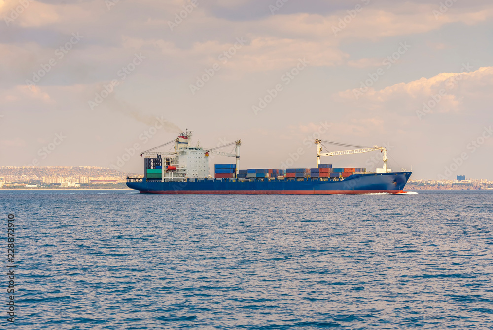Fototapeta premium the ship is a container ship sailing along the sea, against the background of the coastline on the horizon and the sky covered with clouds