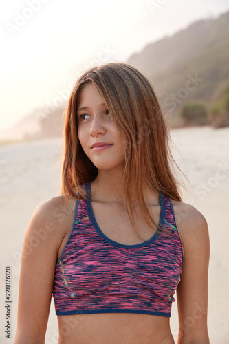 Sporty girl dressed in casual top, has healthy fitness body, feels refreshed after training in open air, looks pensively aside, poses outside, breathes marine air, has active lifestyle, likes sport