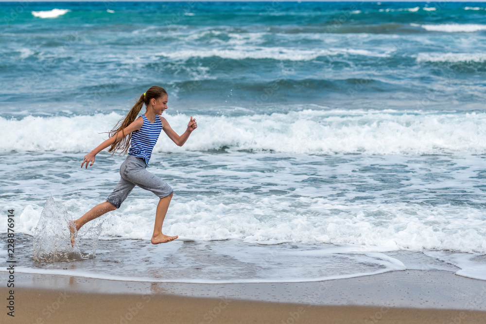 Cute happy little girl running along the beach in jumping over waves. Beautiful summer sunny day, blue sea, picturesque landscape. Cute happy little girl running along the beach in jumping over waves. Beautiful summer sunny day, blue sea, picturesque landscape.