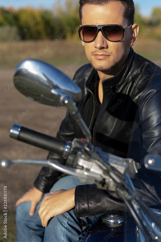 Young man wearing a black leather jacket, sunglasses and jeans sits outdoor on a motorcycle, on a mountain on a blue sky background. Lifestyle, travel. Copy space