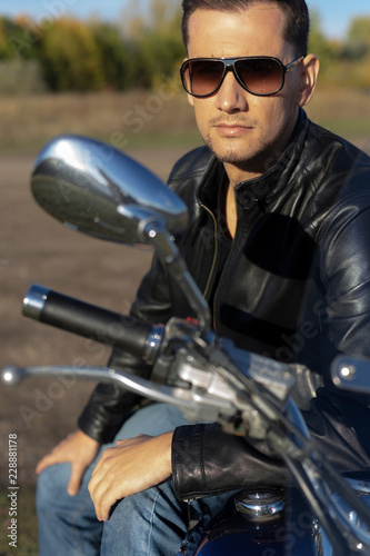 Young man wearing a black leather jacket, sunglasses and jeans sits outdoor on a motorcycle, on a mountain on a blue sky background. Lifestyle, travel. Copy space