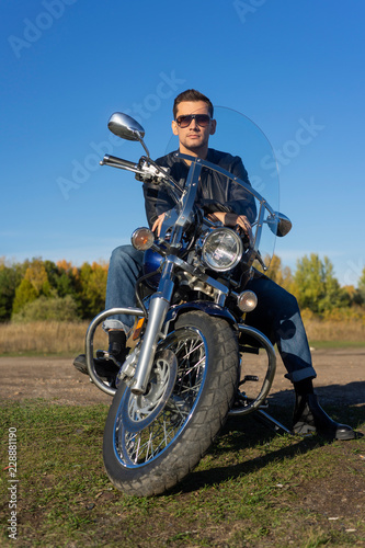 Young man wearing a black leather jacket  sunglasses and jeans sits outdoor on a motorcycle  on a mountain on a blue sky background. Lifestyle  travel. Copy space