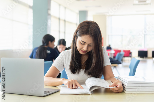 Young asian female student college is reading book while sitting on table with laptop.