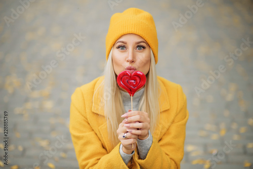 Fashion portrait pretty sweet young woman in yellow coat having fun with red lollipop heart over autumn street background