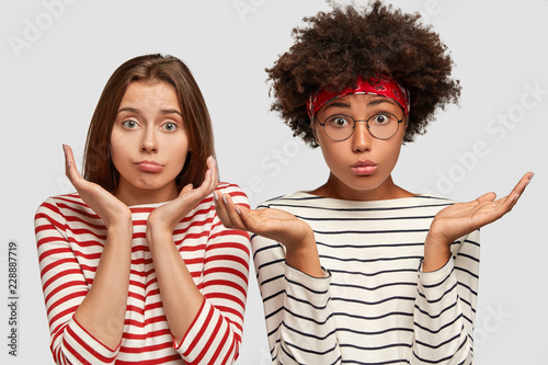 Doubtful two young clueless girls spread palms, have uncertain facial expressions, cant make desicion what topic for project work to choose, wear striped clothes, isolated over white background photo