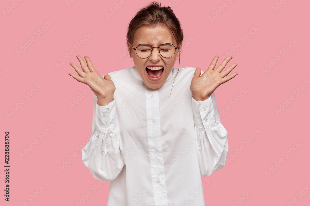 Excited emotional young European woman spreads palms, exclaims very loudly, wears round transparent glasses, dressed in white shirt, poses against pink background. People and emotions concept Excited emotional young European woman spreads palms, exclaims very loudly, wears round transparent glasses, dressed in white shirt, poses against pink background. People and emotions concept