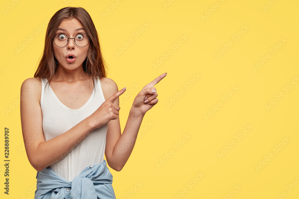 Studio shot of thrilled young lovely female model points witth both index fingers at upper right corner, discusses amazed advertisement, drops jaw with amazement, isolated over yellow studio wall Studio shot of thrilled young lovely female model points witth both index fingers at upper right corner, discusses amazed advertisement, drops jaw with amazement, isolated over yellow studio wall