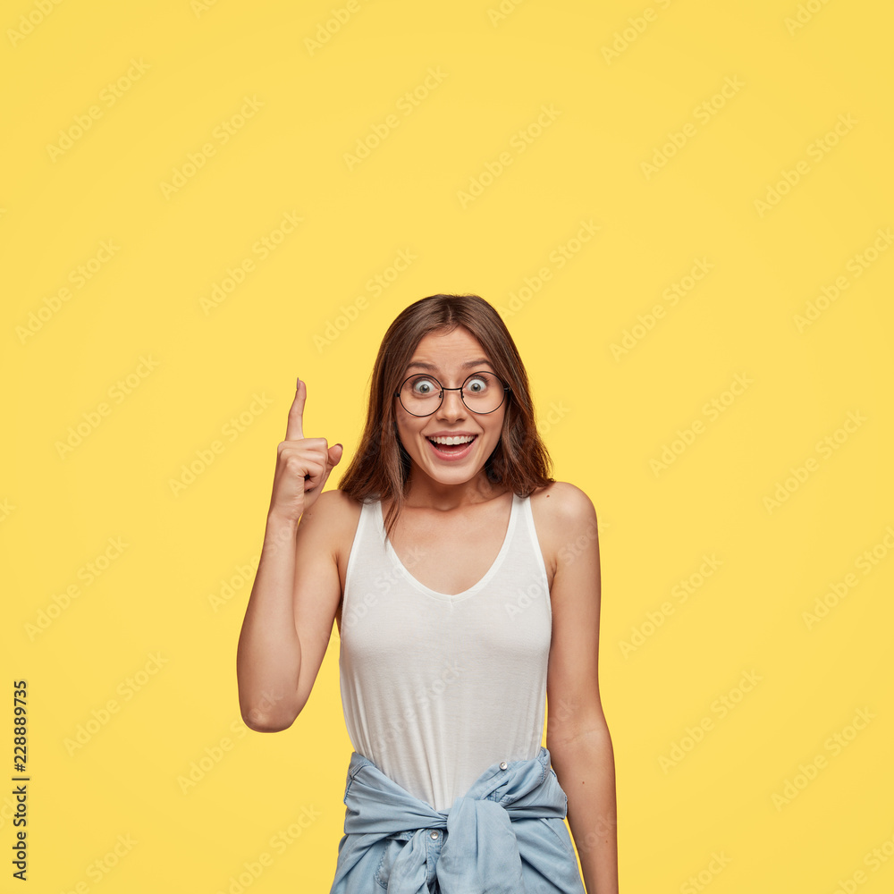 Outgoing pleased happy young female model feels impressed by something upwards, indicates with index finger, wears transparent glasses, dressed in casual clothes, expresses positive emotions Outgoing pleased happy young female model feels impressed by something upwards, indicates with index finger, wears transparent glasses, dressed in casual clothes, expresses positive emotions
