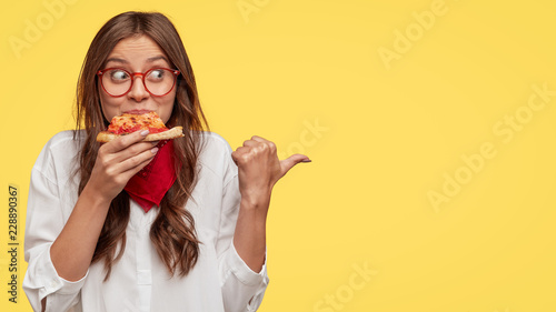 Fotografia Photo of attractive woman eats slice of pizza, points aside with thumb, dressed in fashionable clothes, shows where pizzeria is, isolated over yellow background