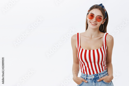 Waist-up shot of stylish attractive european female fashion blogger in trendy round sunglasses headband and striped top holding hands in pockets and smiling friendly and happy at camera