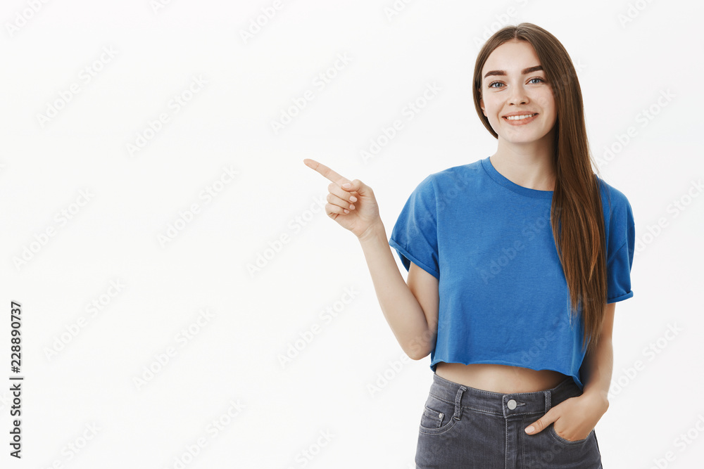 Friendly outgoing beautiful young brunette in blue t-shirt holding hand in pocket of jeans pointing left and smiling cute and polite at camera as if showing way to customer or promoting something Friendly outgoing beautiful young brunette in blue t-shirt holding hand in pocket of jeans pointing left and smiling cute and polite at camera as if showing way to customer or promoting something