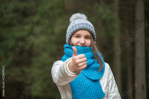 A happy teenager girl is wearing a blue knitted scarf and a gray hat with a pompon, stands against the background of the forest © Goncharenya Tanya