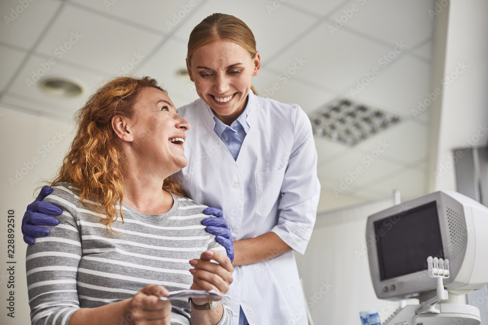 Happiest day. Waist up portrait of joyful mature lady holding ultrasound baby picture while looking at doctor with smile Happiest day. Waist up portrait of joyful mature lady holding ultrasound baby picture while looking at doctor with smile