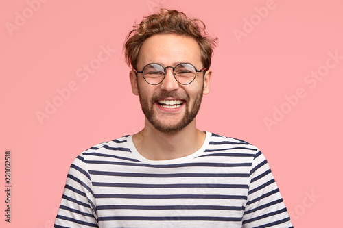 Headshot of pleased hipster has satisfied expression, curly hair and bristle, wears round transparent glasses and striped t shirt, feels glad after promotion at work, isolated over pink background photo
