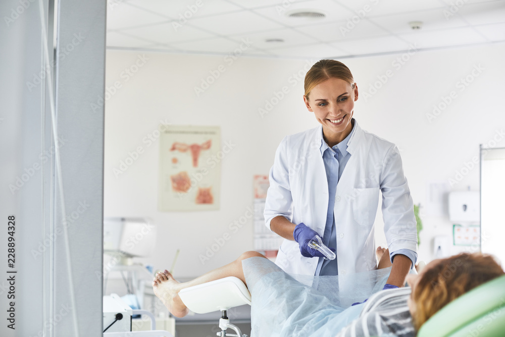 Waist up portrait of gynecologist in white lab coat and sterile gloves using vaginal speculum during pelvic exam. She is looking at patient and smiling Waist up portrait of gynecologist in white lab coat and sterile gloves using vaginal speculum during pelvic exam. She is looking at patient and smiling