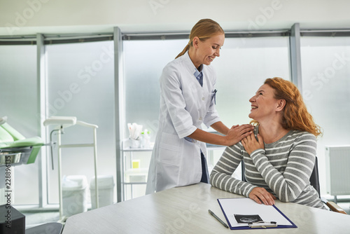 Waist up portrait of joyful gynecologist standing near red-haired lady while she sitting at the table with ultrasound picture of her future baby