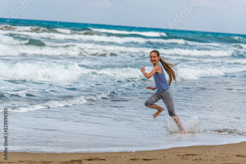 Cute happy Girl in striped t-shirt running along the beach in jumping over waves. Beautiful summer sunny day, blue sea, picturesque landscape.