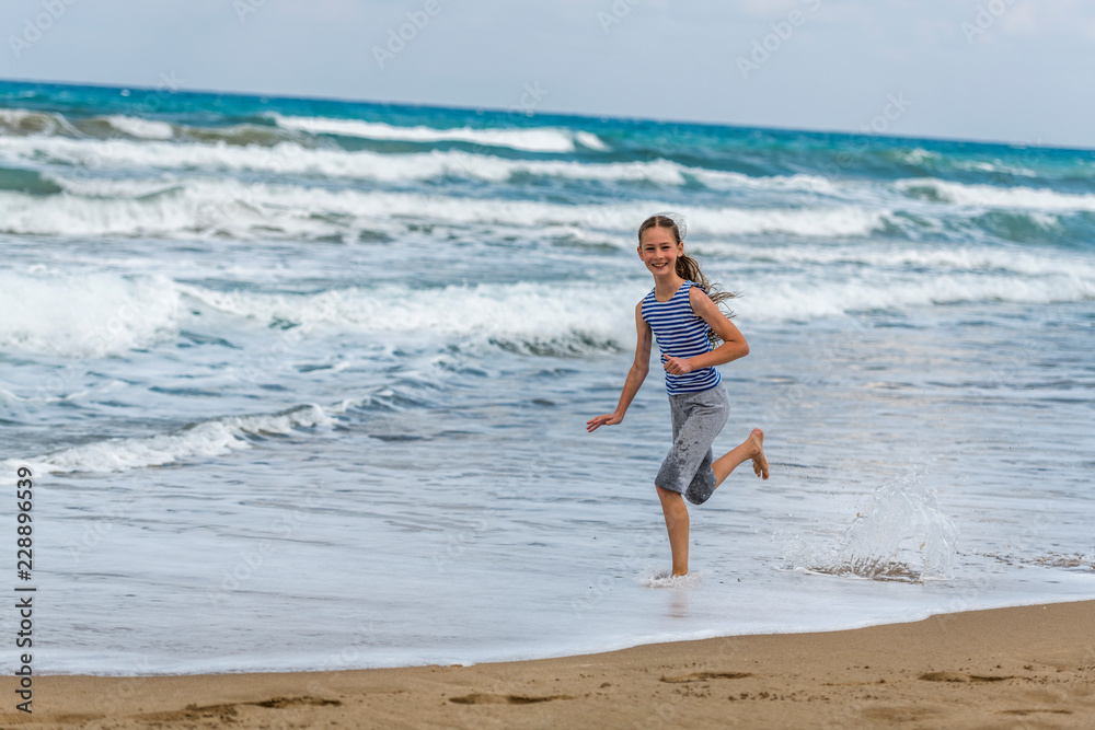 Cute happy Girl in striped t-shirt running along the beach in jumping over waves. Beautiful summer sunny day, blue sea, picturesque landscape. Cute happy Girl in striped t-shirt running along the beach in jumping over waves. Beautiful summer sunny day, blue sea, picturesque landscape.